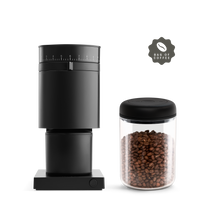 A sleek black grinder from The Grind It Kit by Fellow stands beside a glass jar filled with specialty coffee beans, all set against a solid black backdrop. In the corner, an emblem with a coffee bean icon and the slogan Bag of Coffee is displayed.